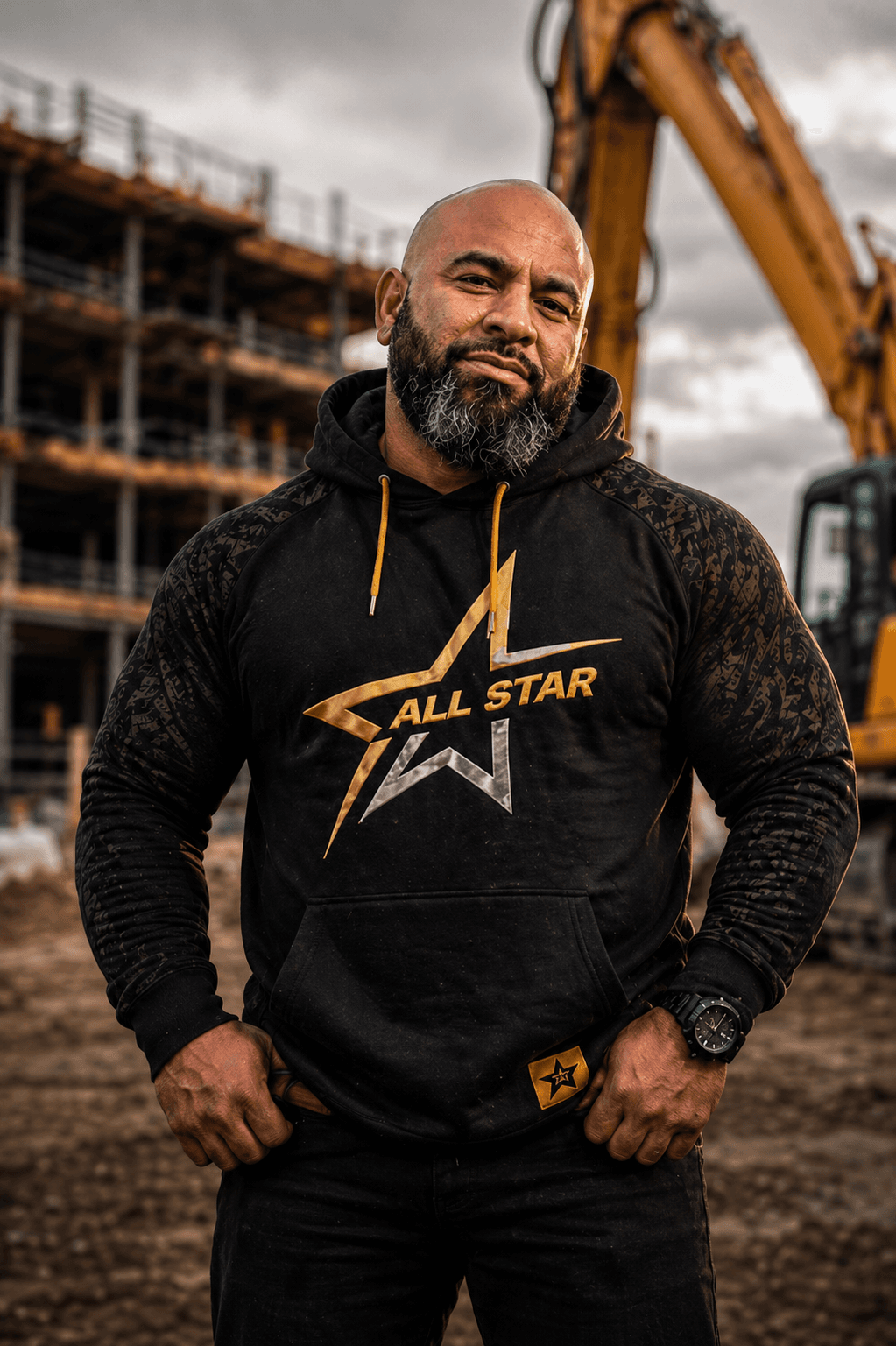 Muscular man in a black All Star hoodie posing at a construction site with excavator.